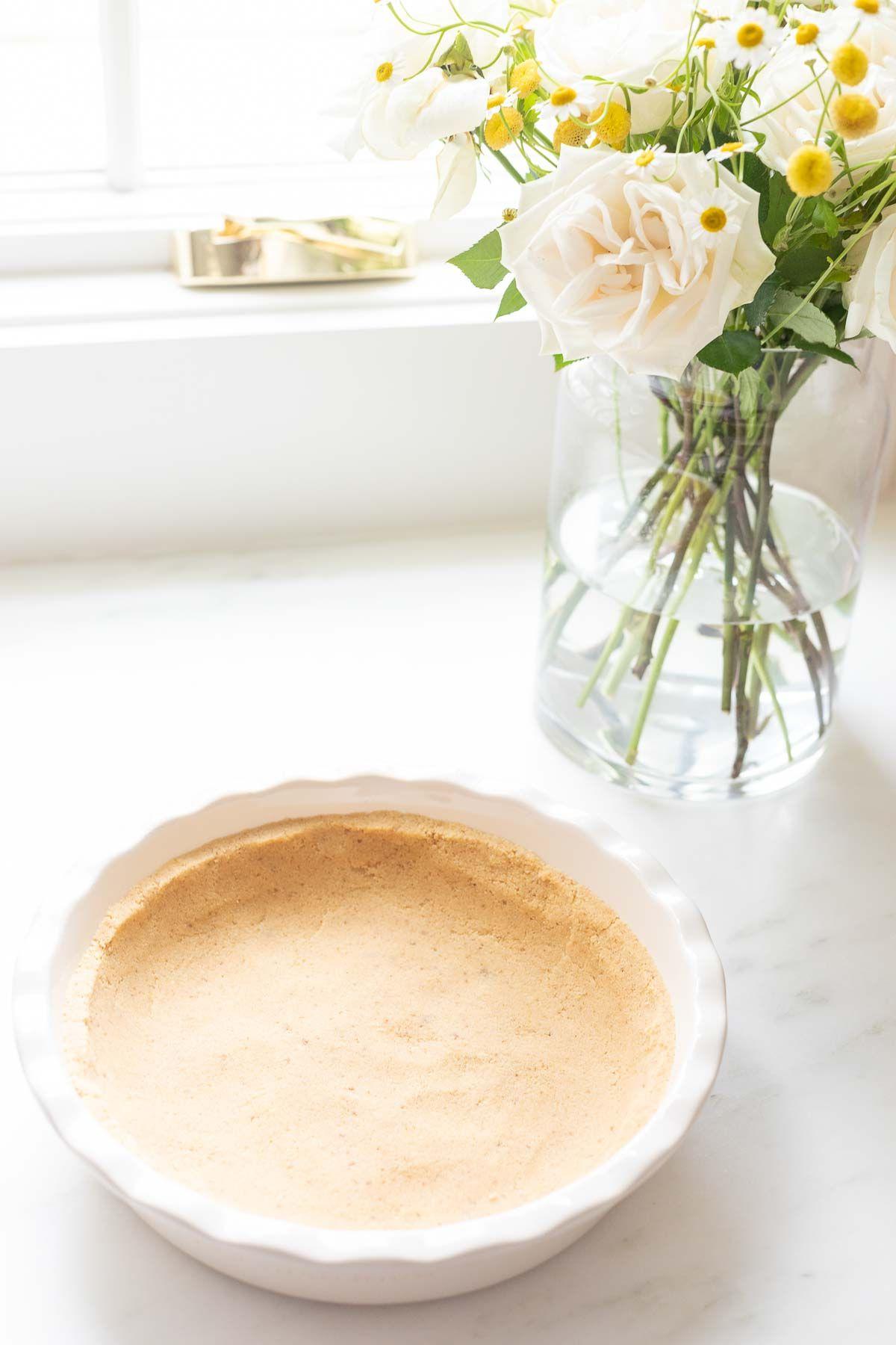 no bake shortbread crust recipe Ein Shortbread-Kuchenboden, in eine weiße, geriffelte Keramik-Kuchenform auf einer Marmoroberfläche gepresst, mit Blumen daneben