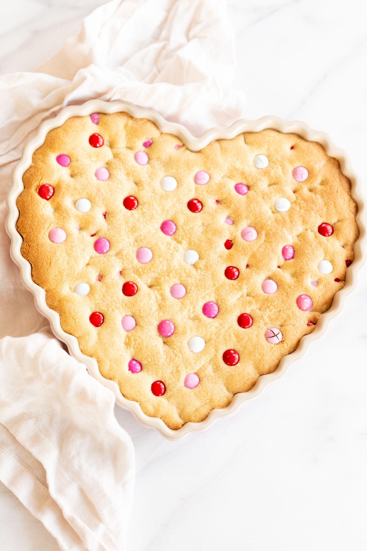 Dicker, zäher M&M-Kekskuchen 2 Dicker und zäher M&M Kekskuchen in einer herzförmigen Form, mit Valentinstags-M&Ms belegt. Eine Leinen-Serviette liegt daneben.