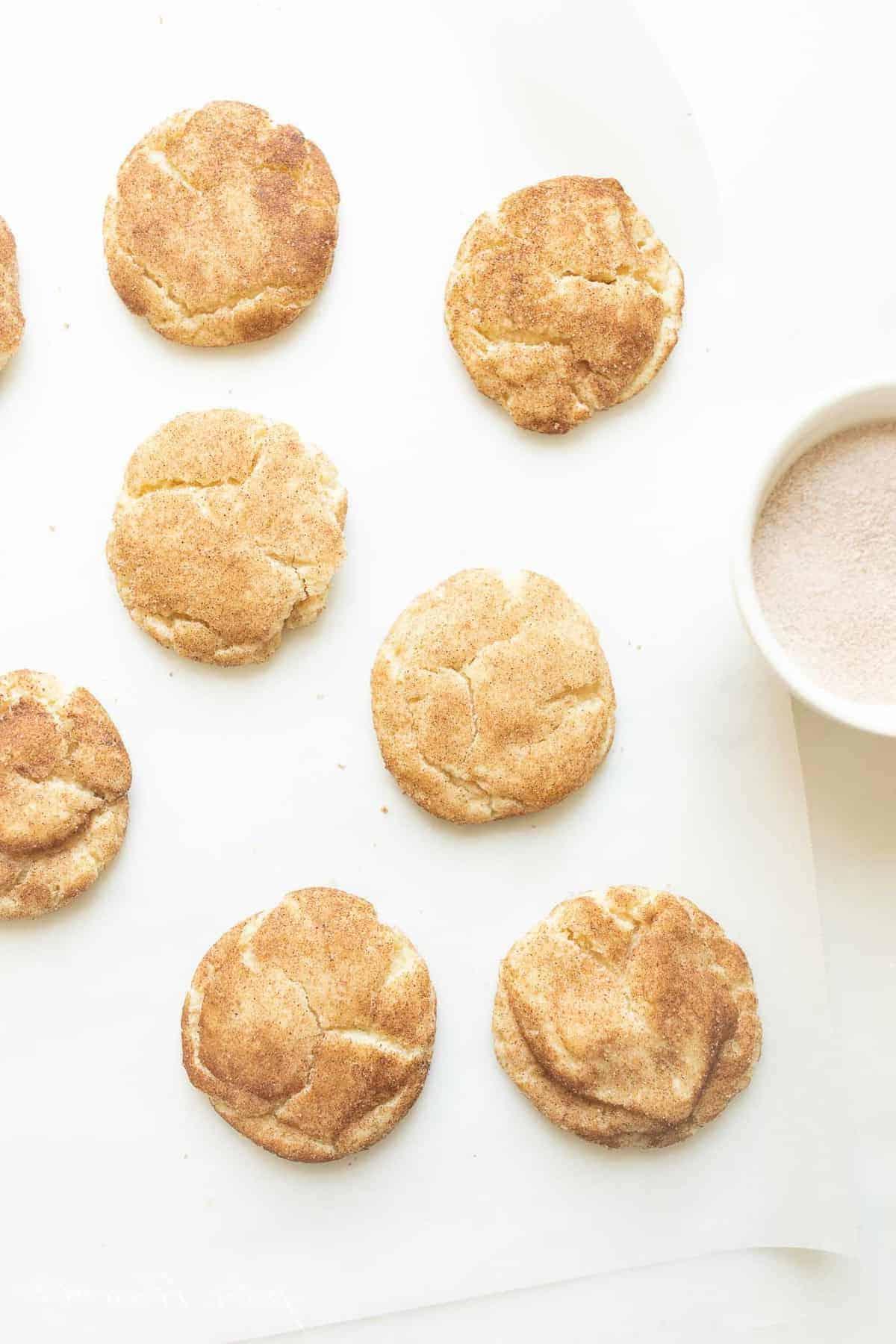 Die besten weichen Snickerdoodles 2 Snickerdoodle-Kekse auf einer weißen Oberfläche mit einer Schüssel Zimt-Zucker daneben.