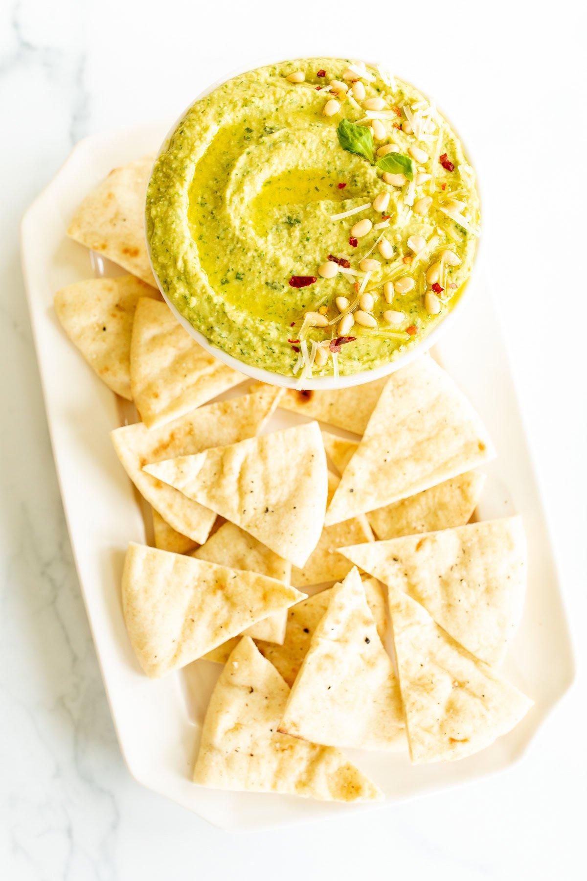 Rezepte mit Pesto 2 Eine Schale grüner Hummus, mit Pesto vermischt und mit Pinienkernen sowie Mikrogrün garniert, wird auf einem rechteckigen weißen Teller mit dreieckigen Fladenbrotstücken serviert.