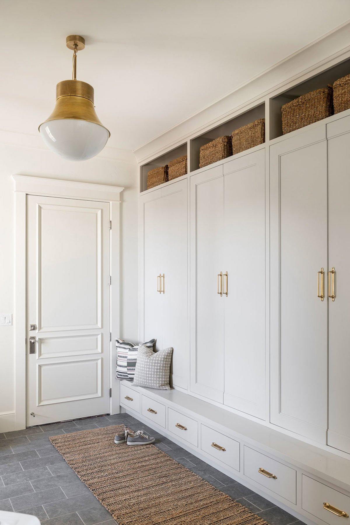 Benjamin Moore Revere Pewter 2 Ein weißer Schlammraum mit Einbauschränken, Weidenkörben oben, einer Bank mit Kissen und einem Teppich auf dem Boden. Die Wände sind in Benjamin Moore Revere Pewter gestrichen. Eine Messing-Deckenleuchte hängt von der Decke.