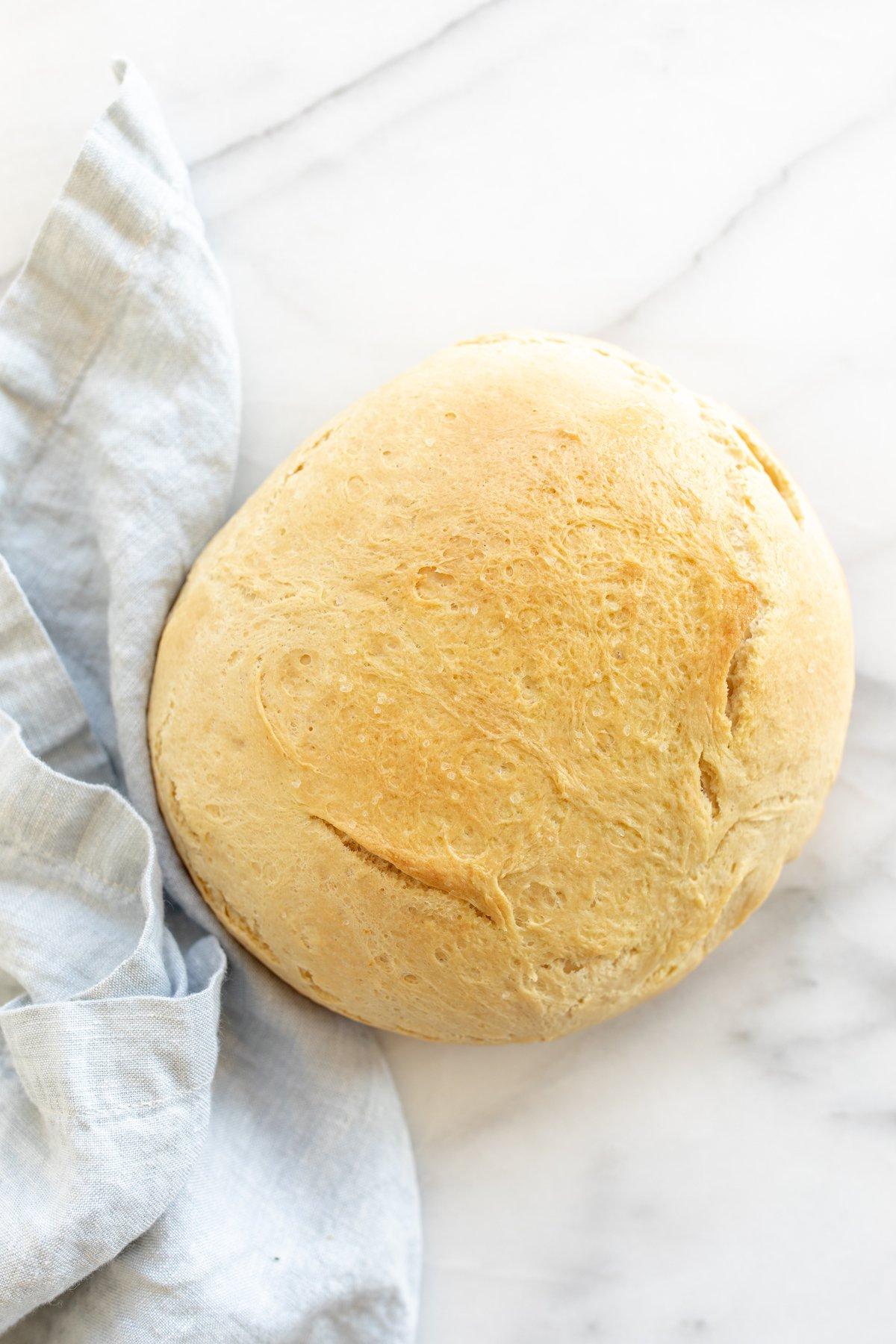 Hausgemachtes Brot im Dutch Oven 2 Eine Marmoroberfläche mit einem runden Laib hausgemachtem Brot aus dem Dutch Oven, daneben eine blaue Leinentischdecke.