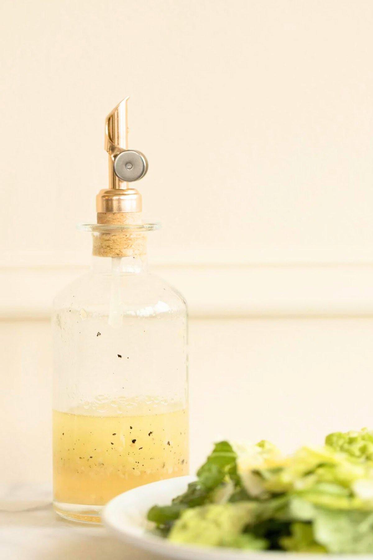Weißwein-Vinaigrette 2 Eine Glasflasche mit einem metallischen Ausgießer enthält eine Weißweinessig-Vinaigrette. Eine Schüssel mit frischem Kopfsalat ist im Vordergrund auf der linken Seite zu sehen.