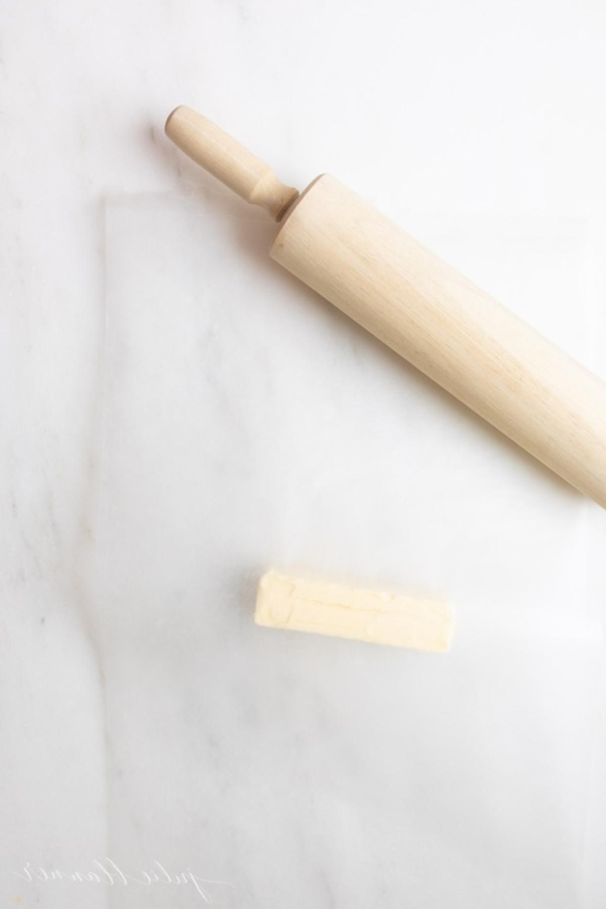 Ein hölzernes Nudelholz neben einem Stück Butter, das zwischen zwei Blättern Wachspapier gelegt ist.