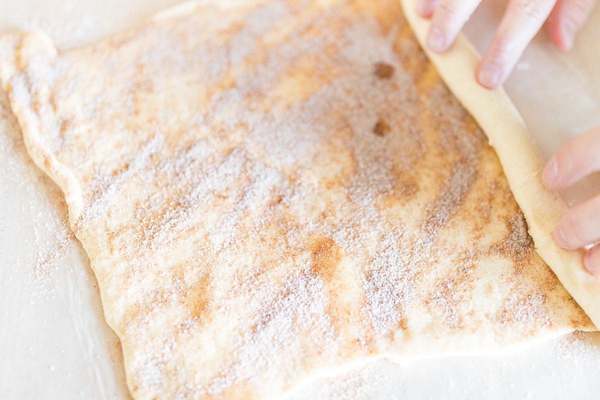 Eine Person bereitet Pekannussbrötchen mit Zimtzucker auf einem Stück Teig zu.