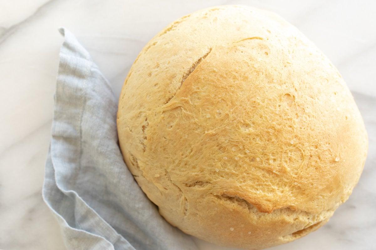 Hausgemachtes Brot im Dutch Oven 11 Eine Marmoroberfläche mit einem runden Laib hausgemachtem Brot, daneben eine blaue Leinentischdecke.