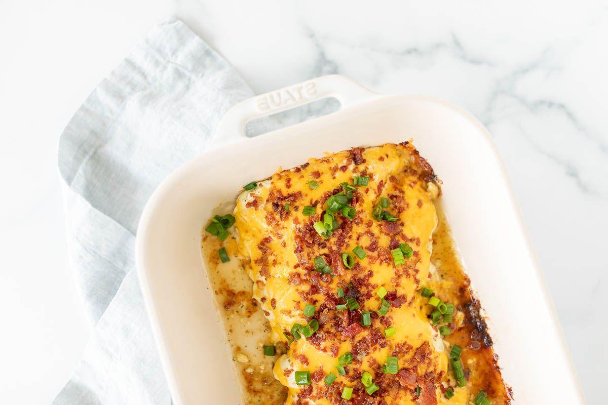 Eine weiße Auflaufform mit cremigem, käsigen 'Crack Chicken', garniert mit Frühlingszwiebeln und gehacktem Speck.