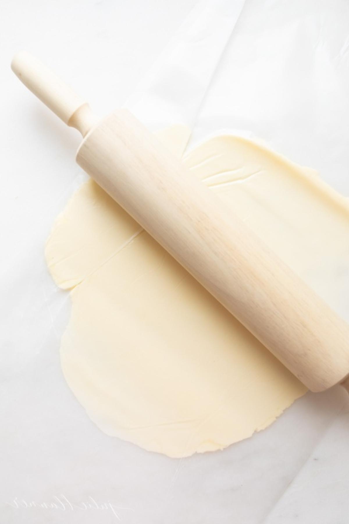 Ein hölzernes Nudelholz neben einem Stück Butter, das zwischen zwei Blättern Wachspapier flachgedrückt ist.