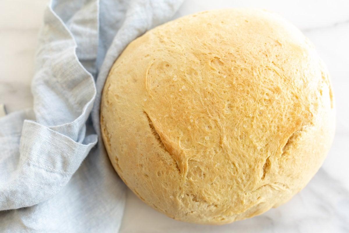 Hausgemachtes Brot im Dutch Oven 12 Eine Marmoroberfläche mit einem runden Laib hausgemachtem Brot, daneben eine blaue Leinentischdecke.