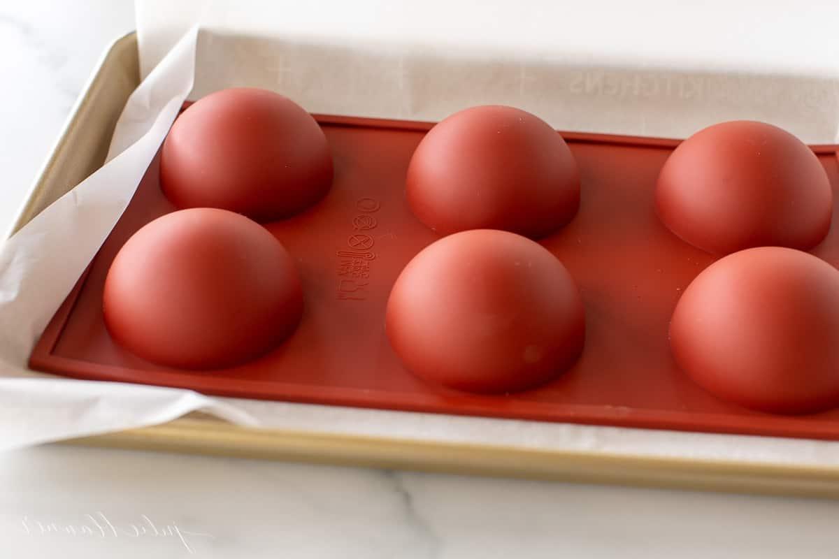 Eine rote Silikonkugelform, gefüllt mit Schokolade für mexikanische Trinkschokolade Bomben, umgedreht auf einem goldenen Backblech.