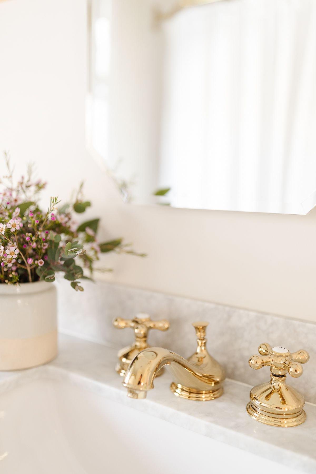 So erstellen Sie ein Spa-Bad zu Hause 12 Ein goldener Wasserhahn auf einem Marmorwaschtisch mit Blumen im Hintergrund, die das elegante Design unterstreichen.