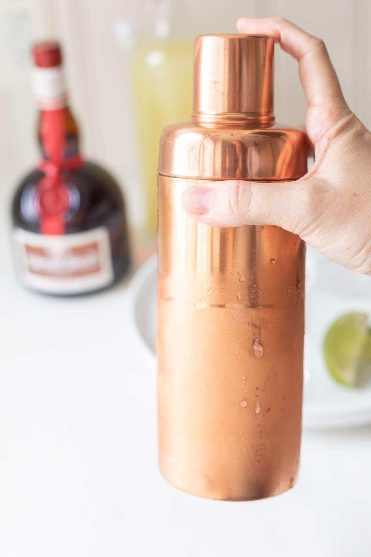 Ein kupferfarbener Shaker mit einer Hand, die Cadillac Margarita Zutaten mixt.