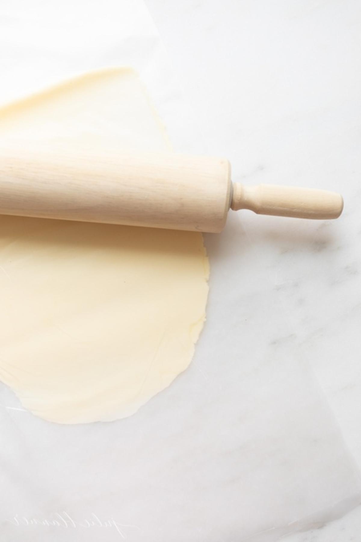 Ein hölzernes Nudelholz neben einem Stück Butter, das zwischen zwei Blättern Wachspapier flachgedrückt ist.