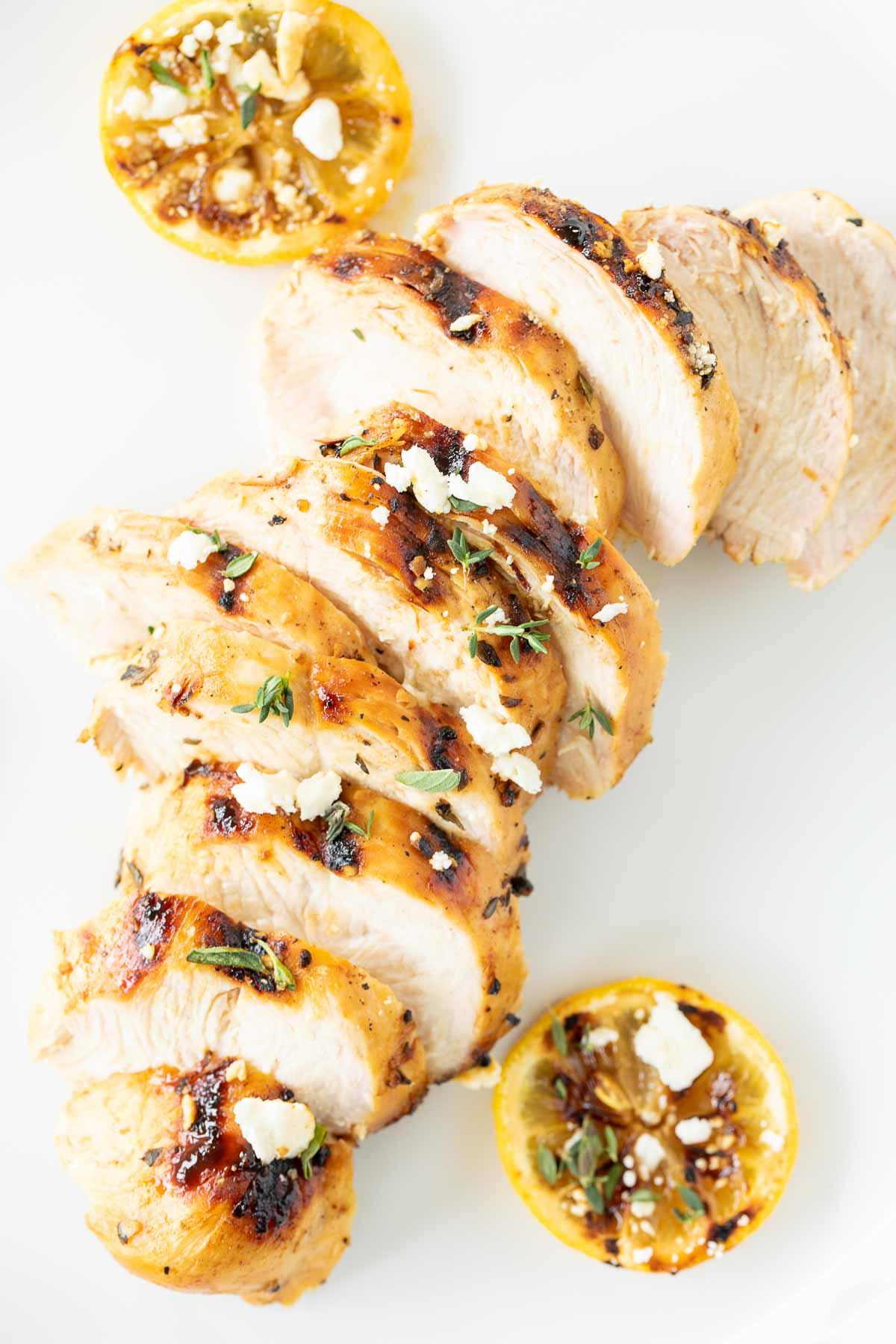 Auf einer weißen Oberfläche liegen Scheiben von griechisch mariniertem Hähnchen, daneben gegrillte Zitrone.