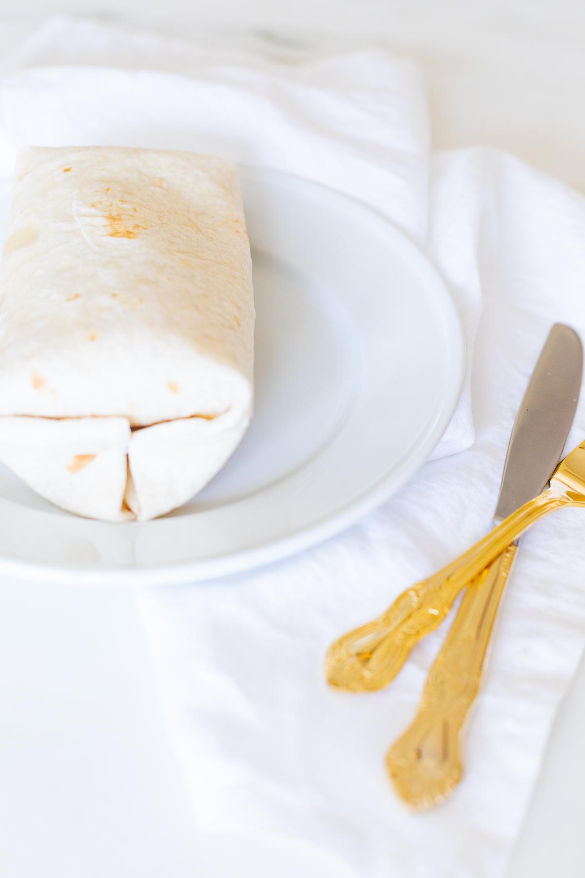 Hähnchen-Wrap-Rezept 14 Ein Hähnchen-Wrap auf einem weißen Teller mit goldenem Besteck.