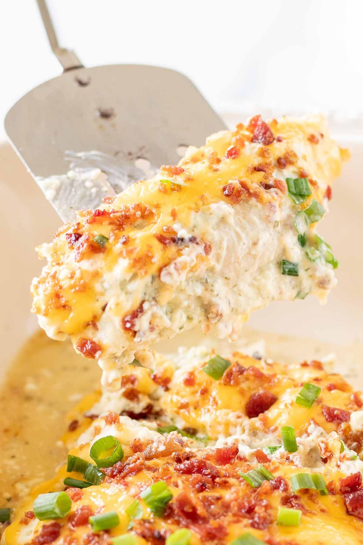 Gebackene 'Crack Chicken'-Hähnchenbrüste in einer weißen Auflaufform, eine silberne Spachtel hebt eine Portion heraus.