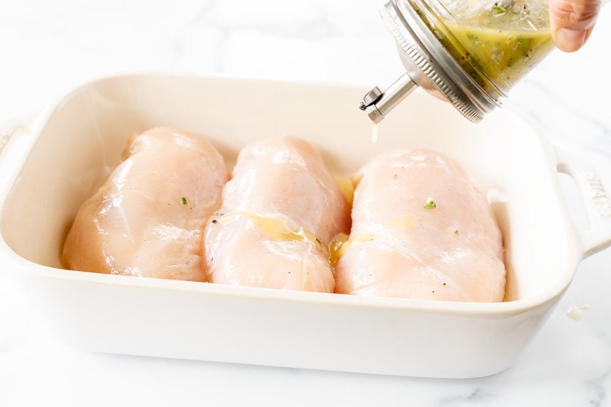 Griechische Hähnchen-Marinade in einem Glasgefäß, wird über Hähnchenbrustfilets in einer weißen Schüssel gegossen.