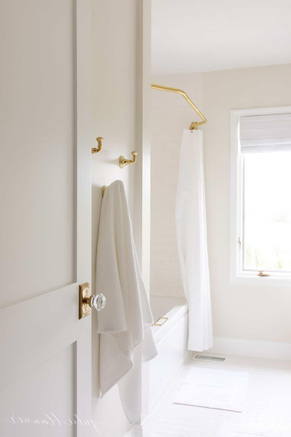 So erstellen Sie ein Spa-Bad zu Hause 15 Ein weißes Badezimmer mit Messing-Handtuchhaken und einer Messing-Duschvorhangstange, die eine warme und organisierte Atmosphäre schaffen.