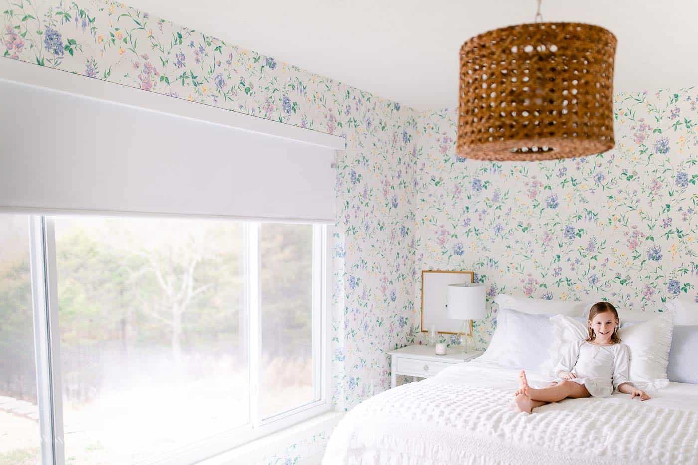 17 Ideen für Kunst über einem Bett 16 Schlafzimmer mit Blumentapete, ein kleines Mädchen liegt auf dem Bett mit einer Fernbedienung für einen motorisierten Rolladen.