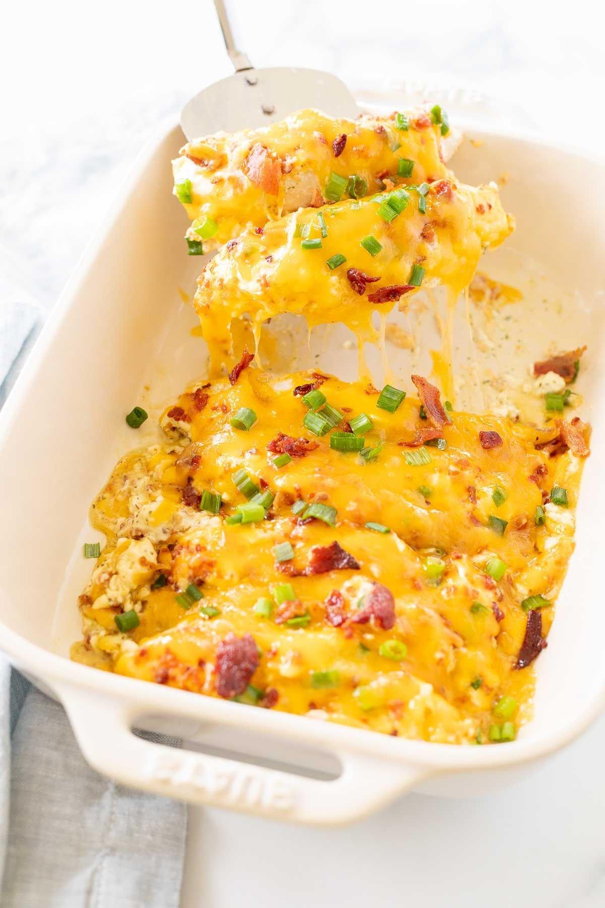 Gebackene 'Crack Chicken'-Hähnchenbrüste in einer weißen Auflaufform, eine silberne Spachtel hebt eine Portion heraus.