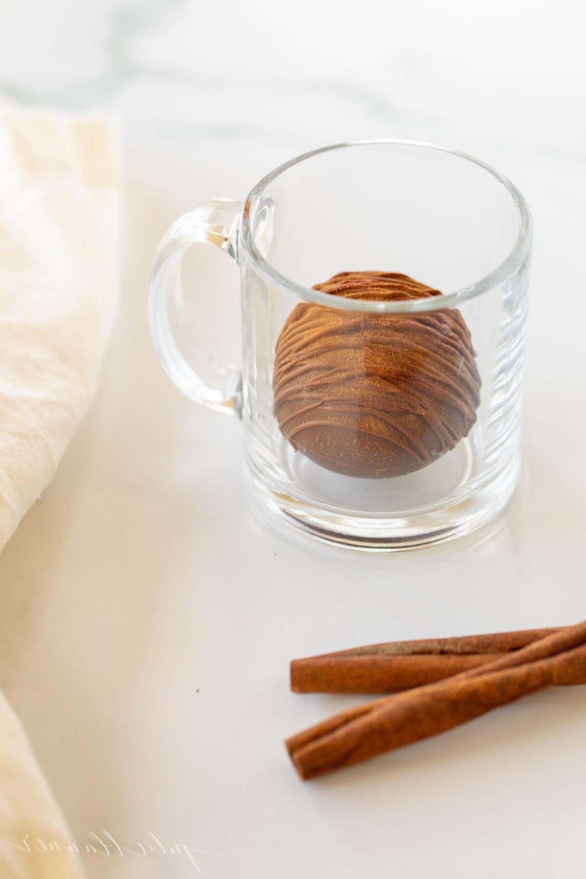 Mexikanische Heiße Schokoladenbomben 19 Eine klare Tasse mit einer mexikanischen Trinkschokolade Bombe, einem Zimtstiel und einer weißen Leinen Serviette daneben, perfekt in Szene gesetzt.