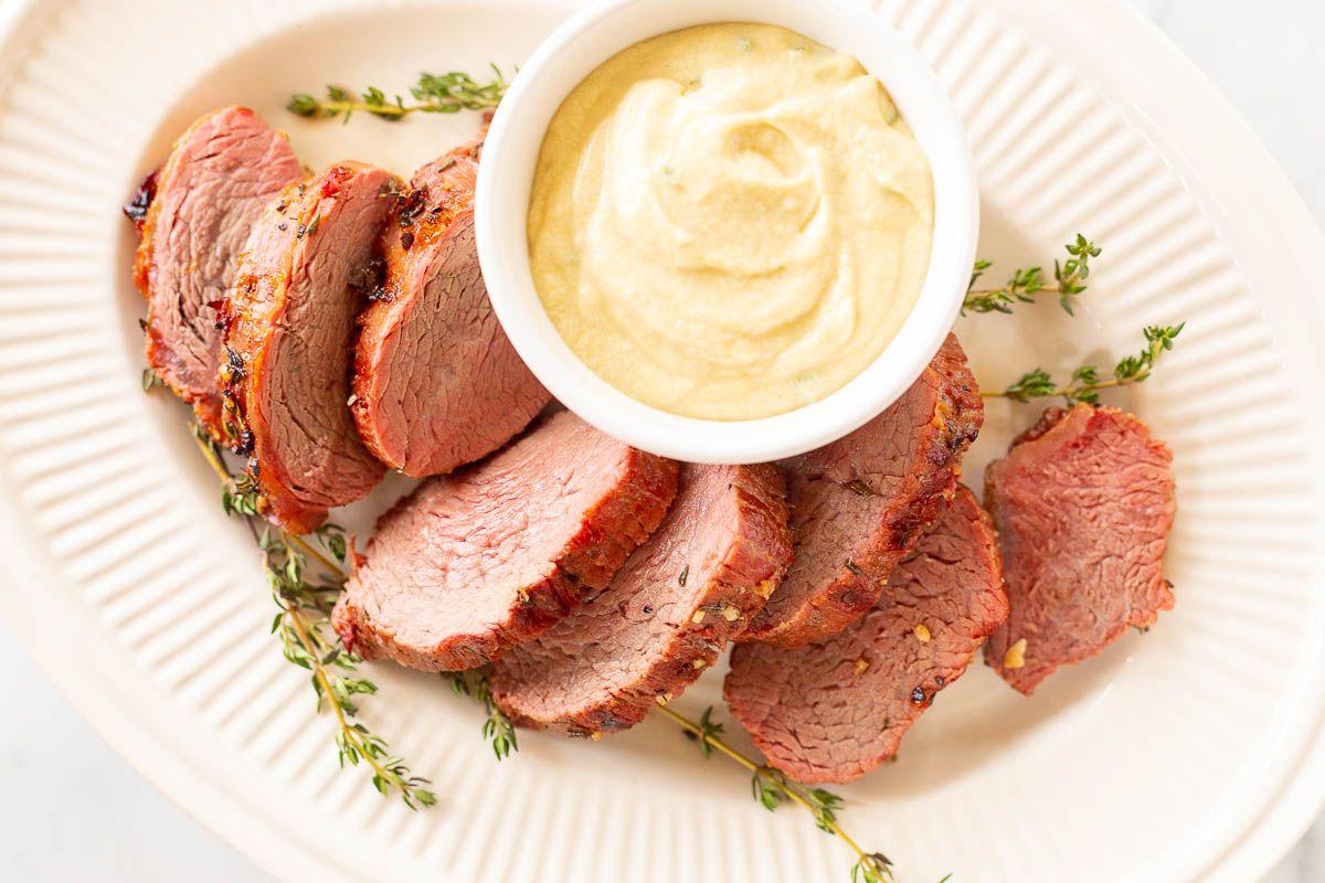 Eine weiße Schale voller Dijon-Senfsauce, umgeben von Rinderfilet auf einer Platte