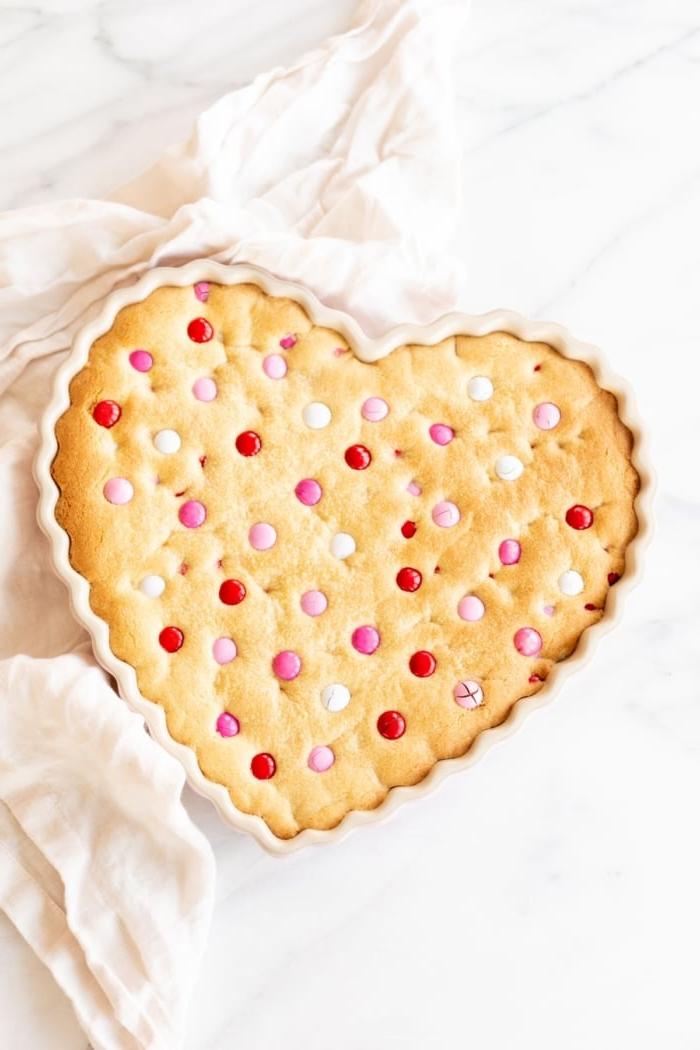 Dicker, zäher M&M-Kekskuchen 3 Dicker und zäher M&M Kekskuchen in einer herzförmigen Form, mit Valentinstags-M&Ms belegt. Eine Leinen-Serviette liegt daneben.