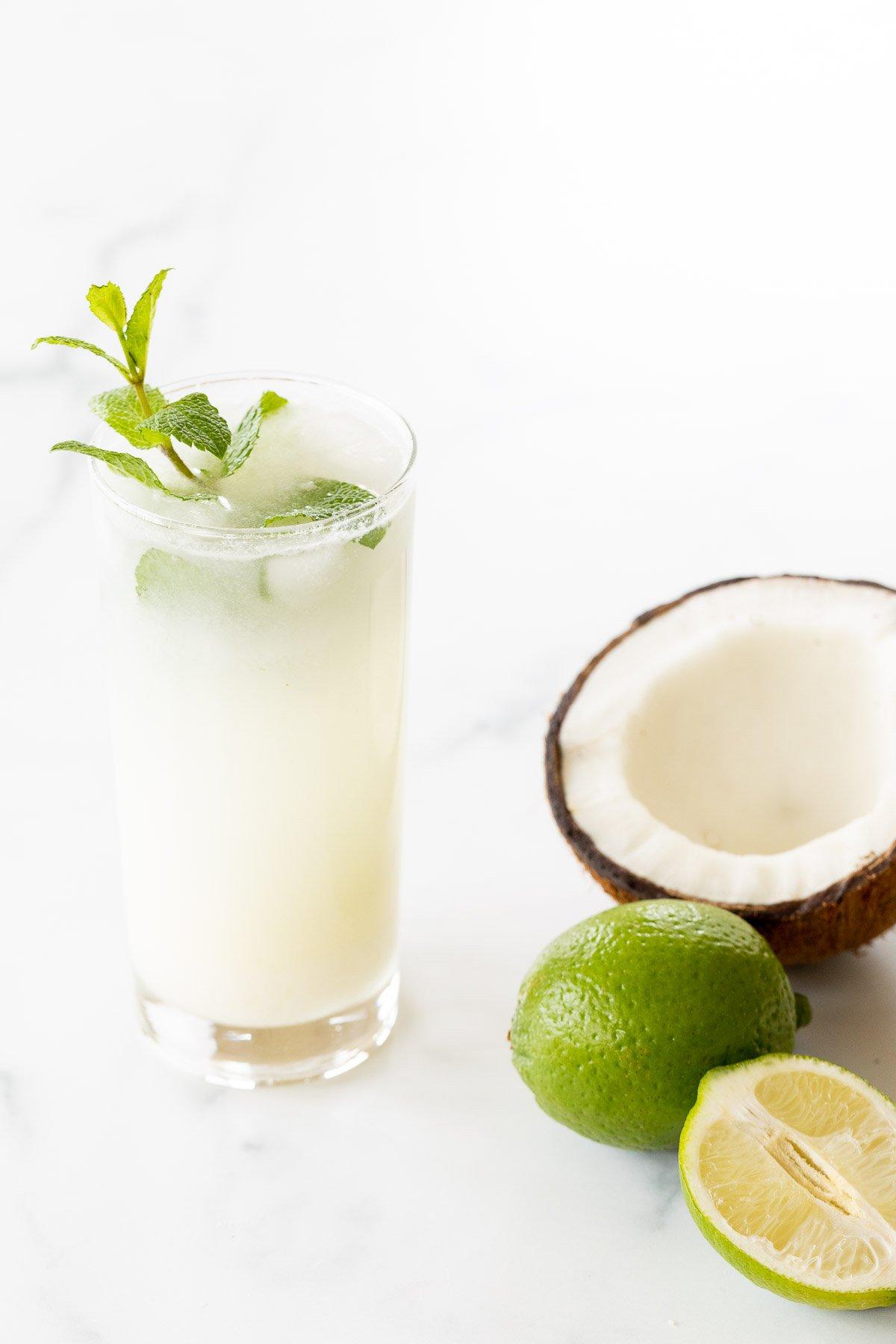 Kokosnuss-Mojito 3 Ein Kokos-Mojito, garniert mit Minze, auf einer Marmorfläche. Limetten und eine offene Kokosnuss daneben.