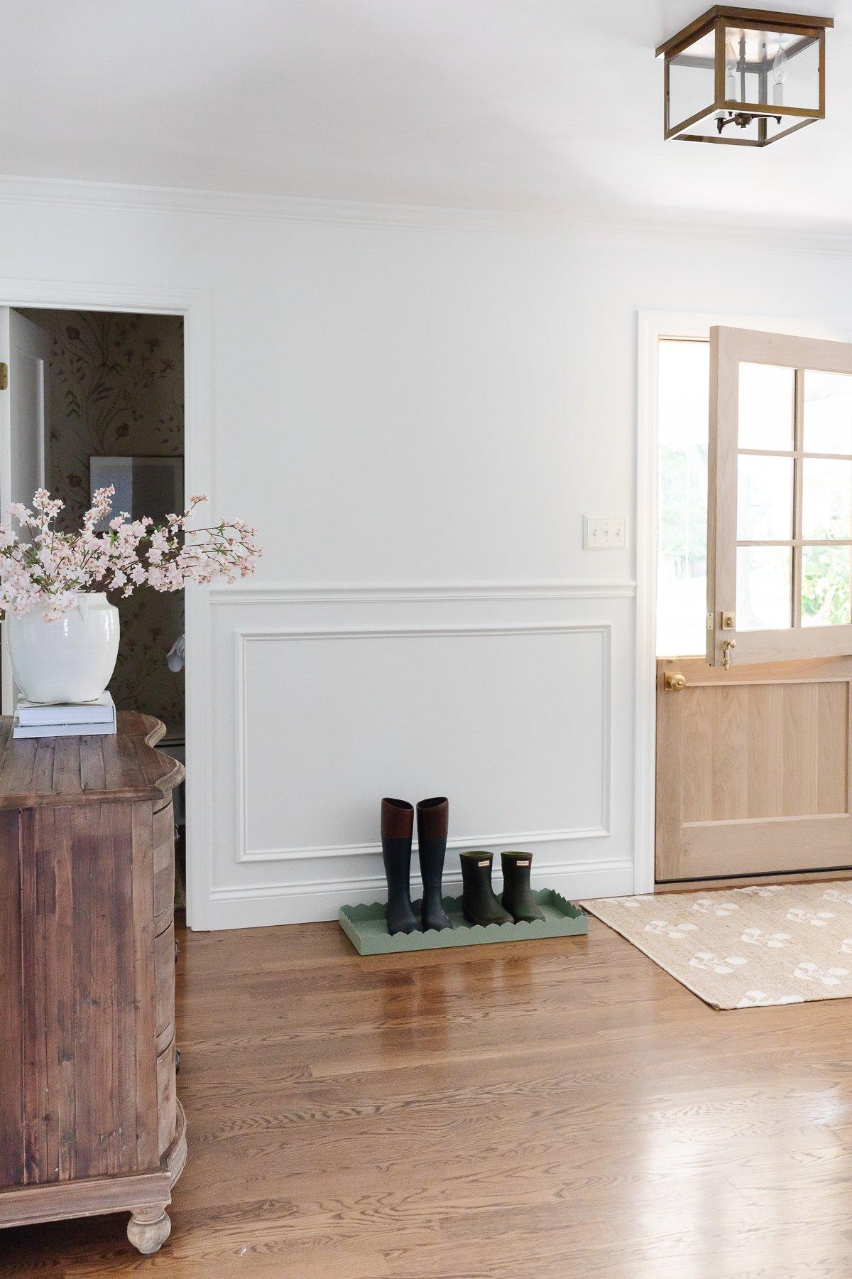 Holzboden-Lasurfarben 6 Ein elegantes weißes Foyer-Design mit einer holländischen Tür, klassischen Kassettenleisten an den Wänden und einem warmen, neutral gebeizten Hartholzboden.