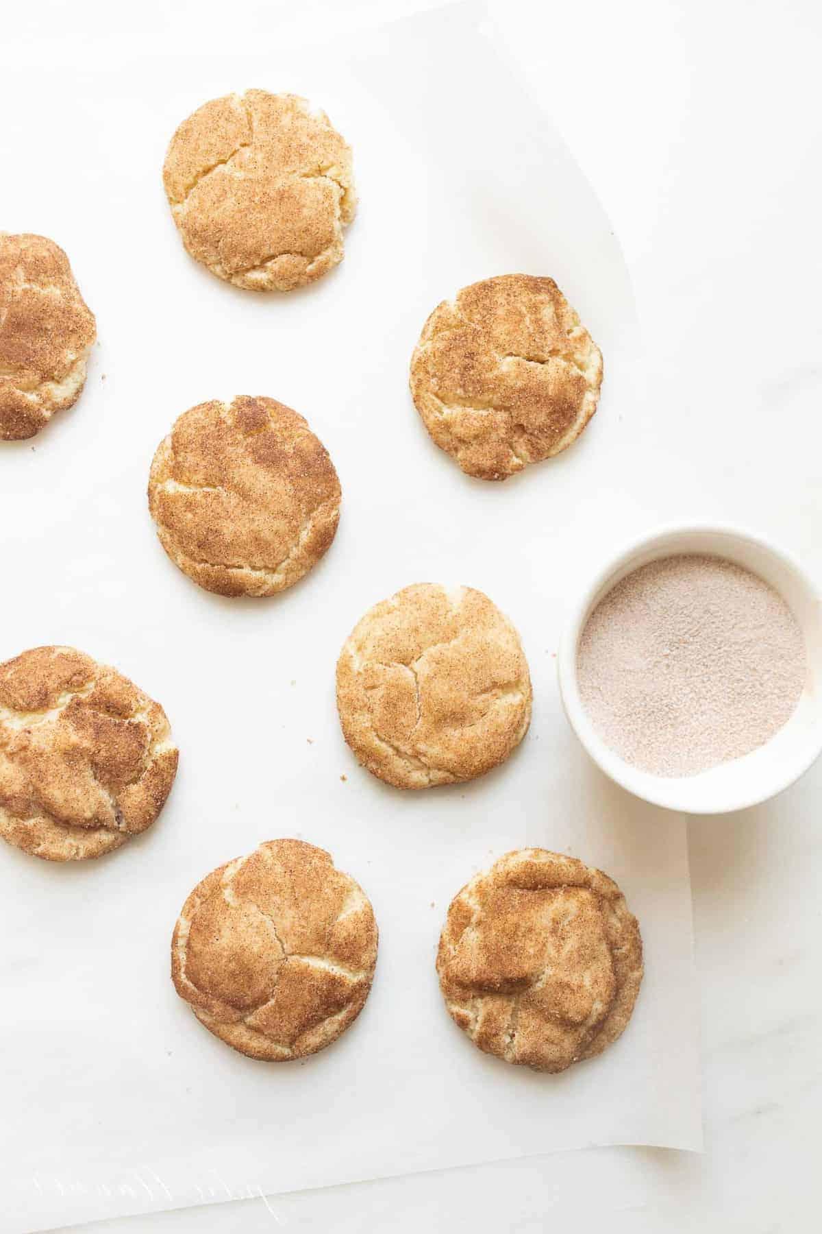 Die besten weichen Snickerdoodles 3 Snickerdoodle-Kekse auf einer weißen Oberfläche mit einer Schüssel Zimt-Zucker daneben.