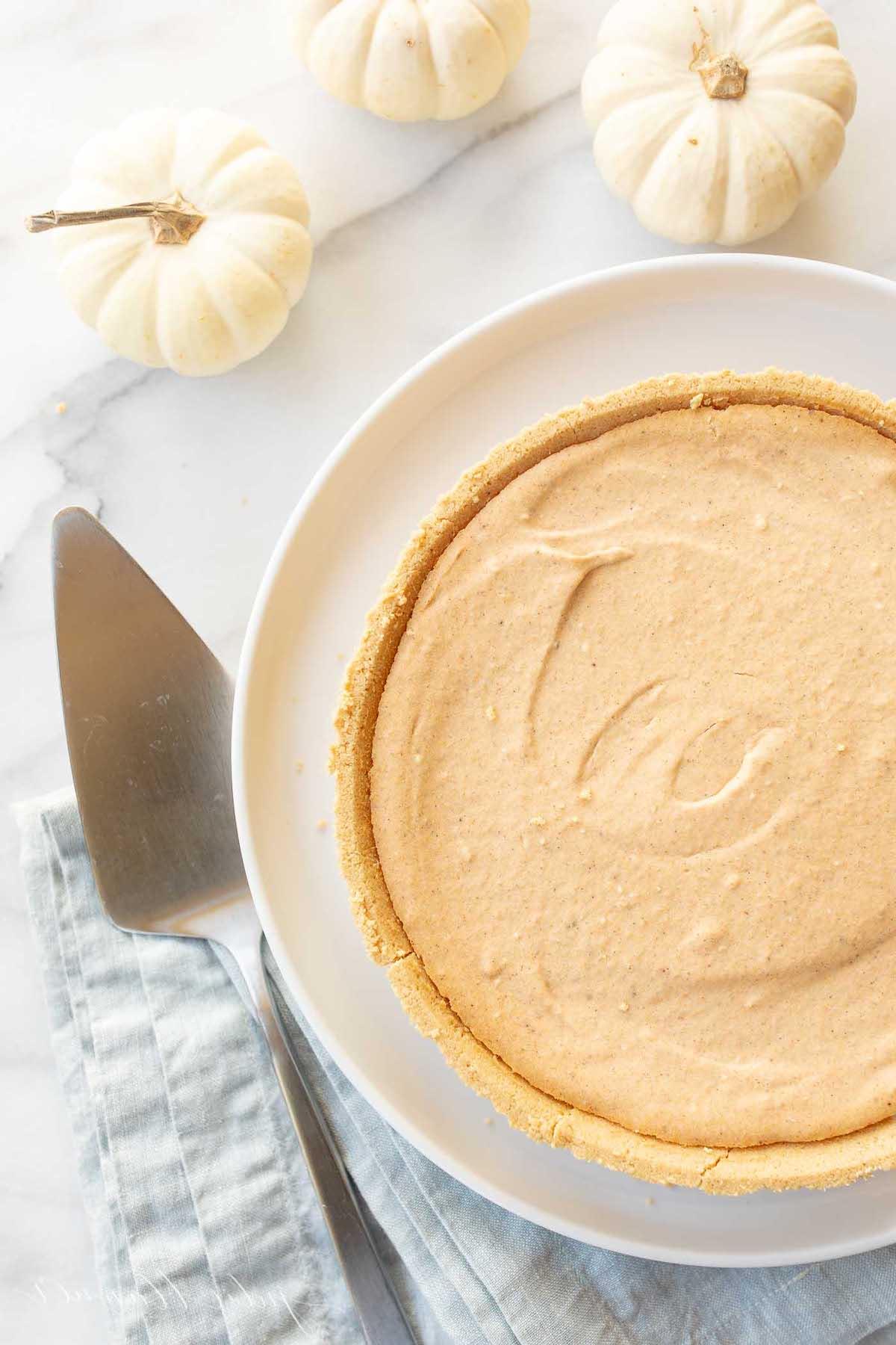 Einfaches Kürbis-Käsekuchen-Rezept ohne Backen 3 Ein No-Bake Kürbis-Käsekuchen auf einem weißen Teller, daneben ein Tortenheber und kleine weiße Kürbisse im Hintergrund.