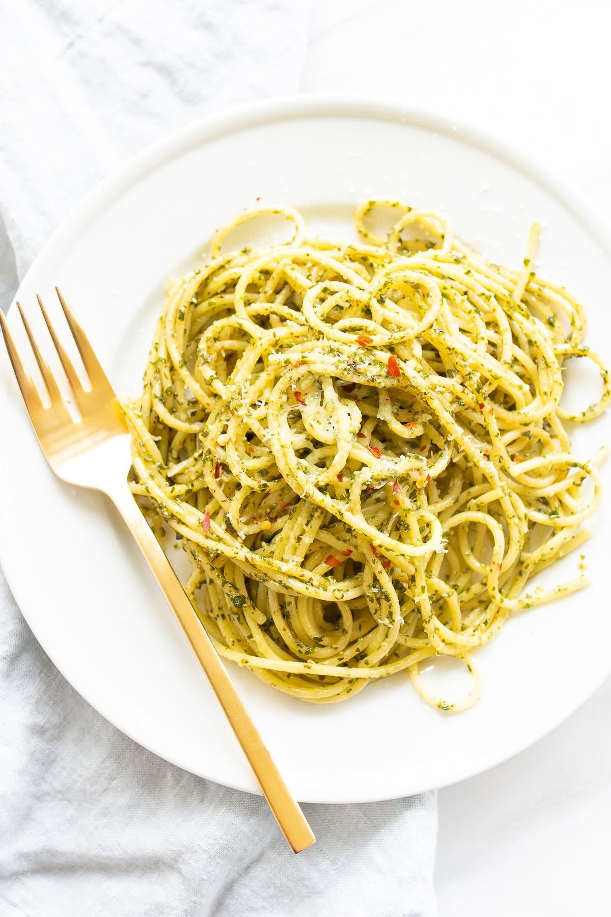 Rezepte mit Pesto 3 Ein weißer Teller mit einer Portion Spaghetti, die in grüner Pesto-Soße geschwenkt und mit roten Pfefferflocken garniert ist. Eine goldene Gabel liegt elegant rechts neben dem Teller. Dieses Gericht ist perfekt für alle, die Pesto-Rezepte erkunden möchten, und bietet ein lebhaftes und geschmackvolles Erlebnis.