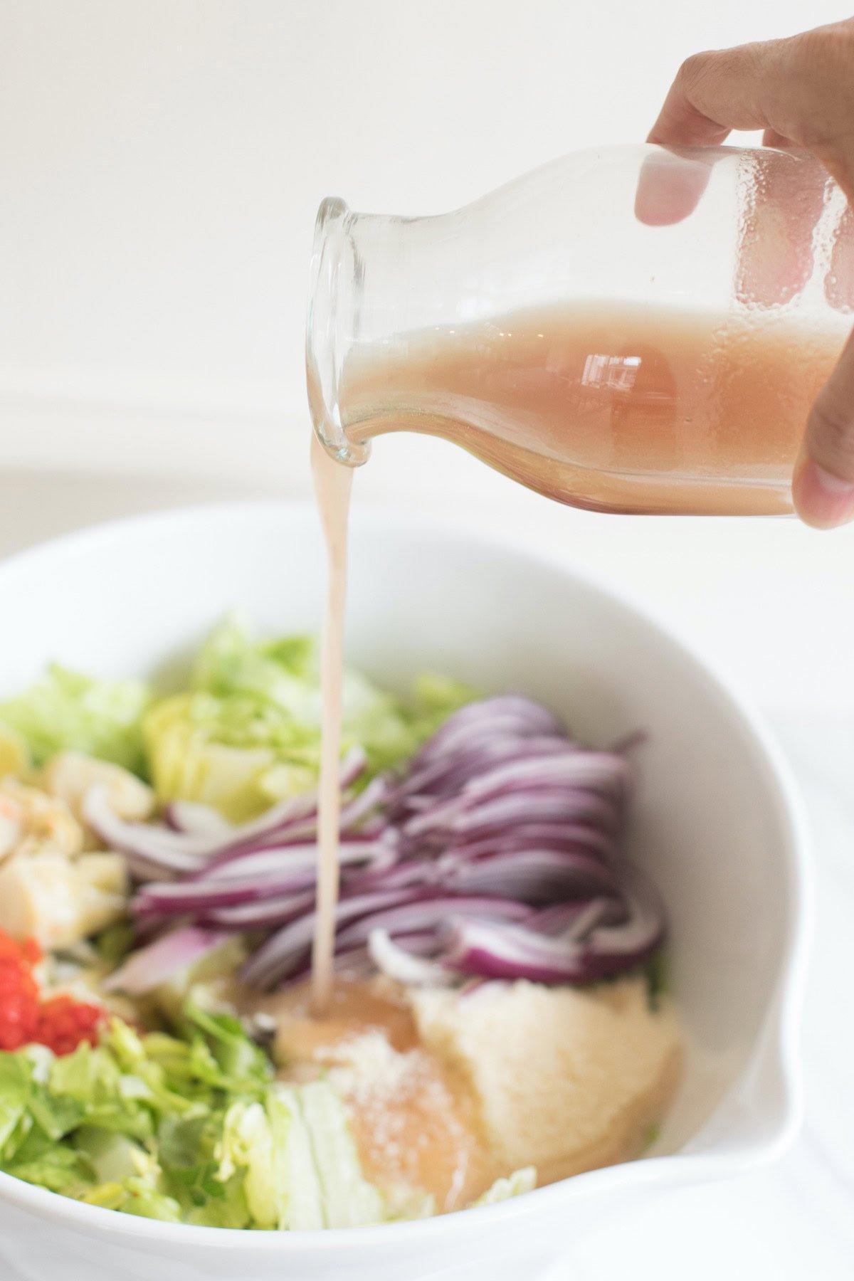 Rotweinessig-Vinaigrette 3 Eine Hand gießt eine hellbraune Rotweinvinaigrette aus einer Glasflasche in eine Schüssel mit gehacktem Salat, geschnittenen roten Zwiebeln und anderen Salatzutaten.