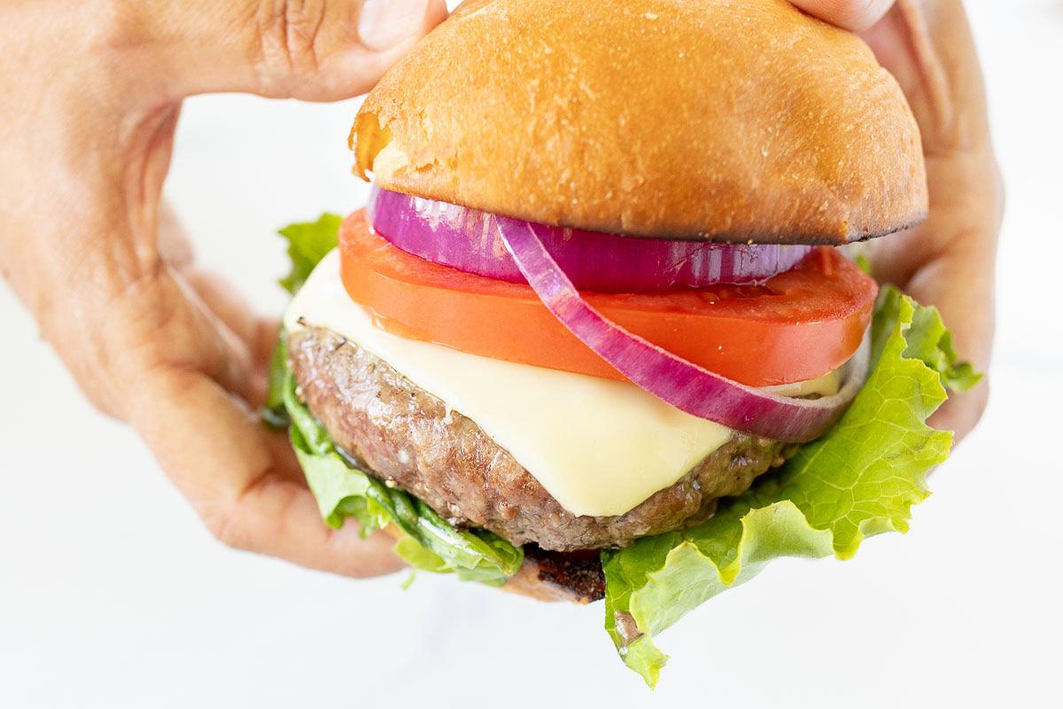 Burger Beilagen 3 Eine Person hält einen Hamburger mit Salat, einem Rindfleisch-Patty, geschmolzenem Käse, Tomatenscheiben, roten Zwiebelringen und einem Brötchen – perfekt kombiniert mit Ihren Lieblings-Burger-Beilagen.