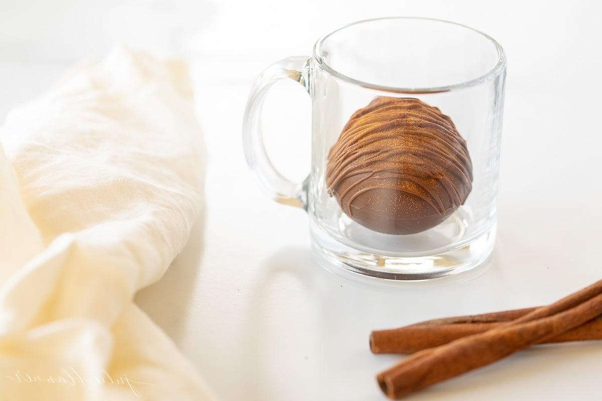 Mexikanische Heiße Schokoladenbomben 3 Eine klare Tasse mit einer mexikanischen Trinkschokolade Bombe, einem Zimtstiel und einer weißen Leinen Serviette daneben.