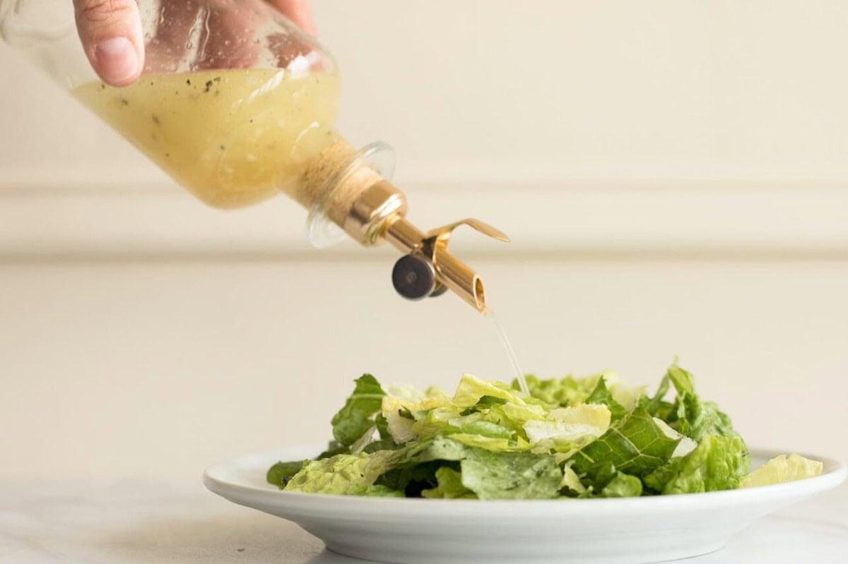 Weißwein-Vinaigrette 3 Eine Hand gießt Weißweinessig-Vinaigrette aus einer Glasflasche auf einen Teller mit grünem Blattsalat, der auf einer weißen Oberfläche steht.