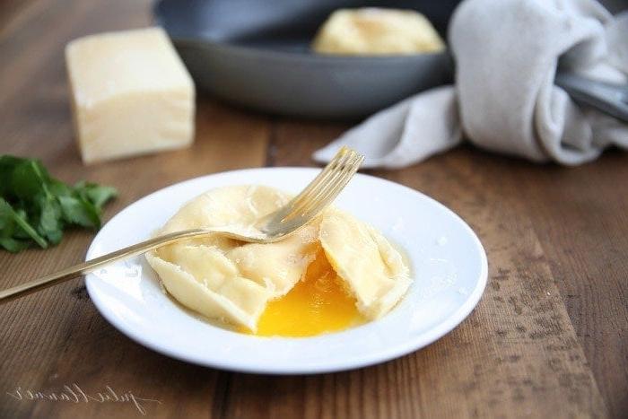 Hausgemachte Eier-Ravioli sind schnell und einfach zuzubereiten und eignen sich hervorragend als Vorspeise oder Hauptgericht. Ravioli gefüllt mit Käse und einem Eigelb, das einfach so beeindruckend ist. Eine angeschnittene Eier-Ravioli auf einem weißen Teller, das flüssige Eigelb läuft heraus, daneben eine Gabel.