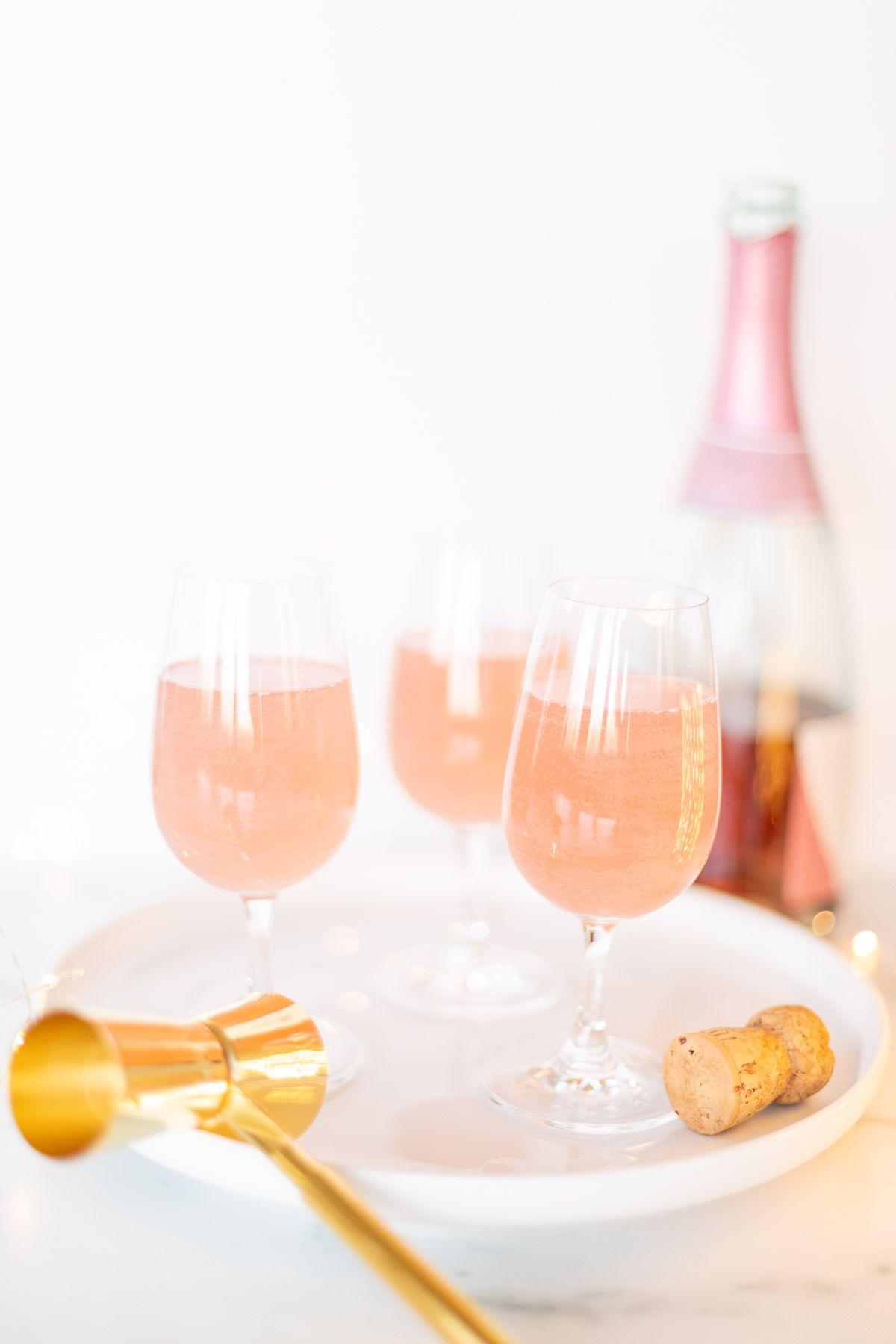 Muttertags-Brunch-Ideen 29 3 Gläser Champagner-Margaritas auf einem weißen Tablett, goldener Konfetti umrandet und eine Flasche pinker Champagner im Hintergrund