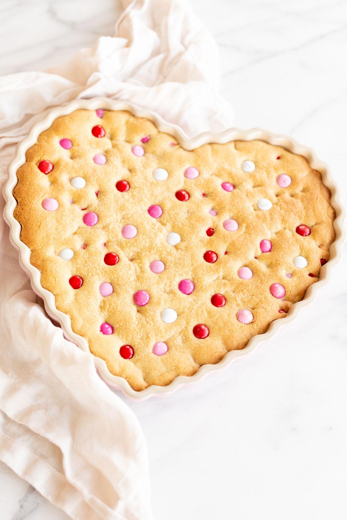 Dicker, zäher M&M-Kekskuchen 4 Dicker und zäher M&M Kekskuchen in einer herzförmigen Form, mit Valentinstags-M&Ms belegt. Eine Leinen-Serviette liegt daneben.
