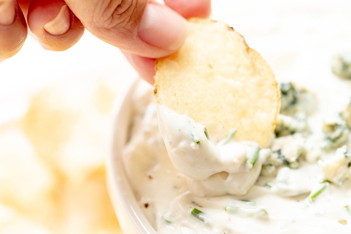 Eine Hand greift einen knusprigen Kartoffelchip, um ihn in den cremigen Blauschimmelkäse-Dip in einer weißen Schale zu tunken – ein verlockender Snackmoment.