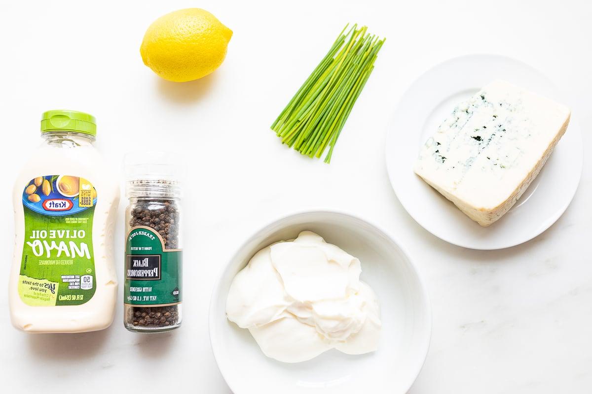 Die frischen Zutaten für den Blauschimmelkäse Dip – Sauerrahm, Mayonnaise, Blauschimmelkäse, Zitrone und Frühlingszwiebeln – appetitlich auf einer weißen Küchenarbeitsplatte arrangiert, bereit zur Zubereitung.