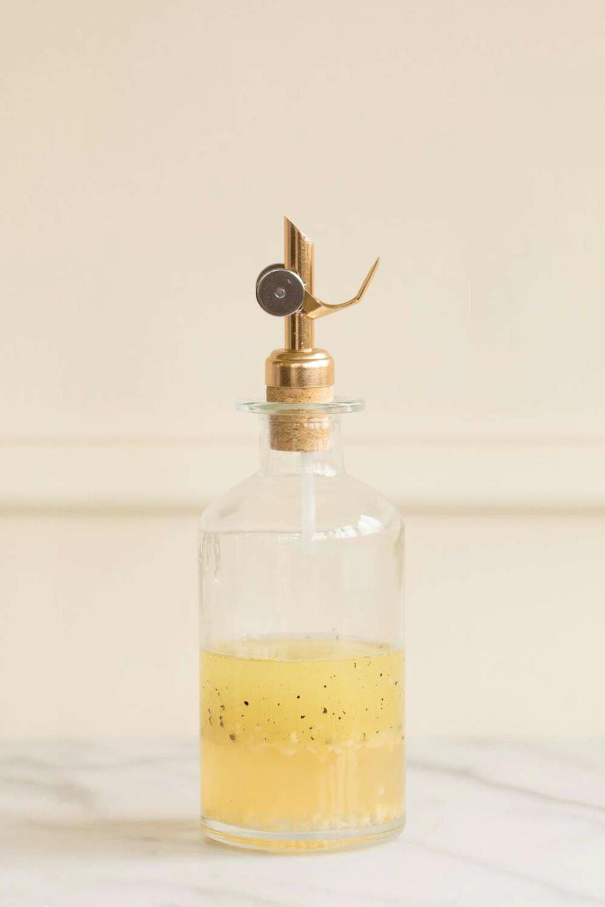 Weißwein-Vinaigrette 5 Klarer Glas-Ölspender mit Metallausgießer, teilweise gefüllt mit einer gelben Flüssigkeit, die schwarze Sprenkel enthält – einer hausgemachten Weißweinessig-Vinaigrette ähnelnd – auf einer weißen Oberfläche vor hellem Hintergrund.