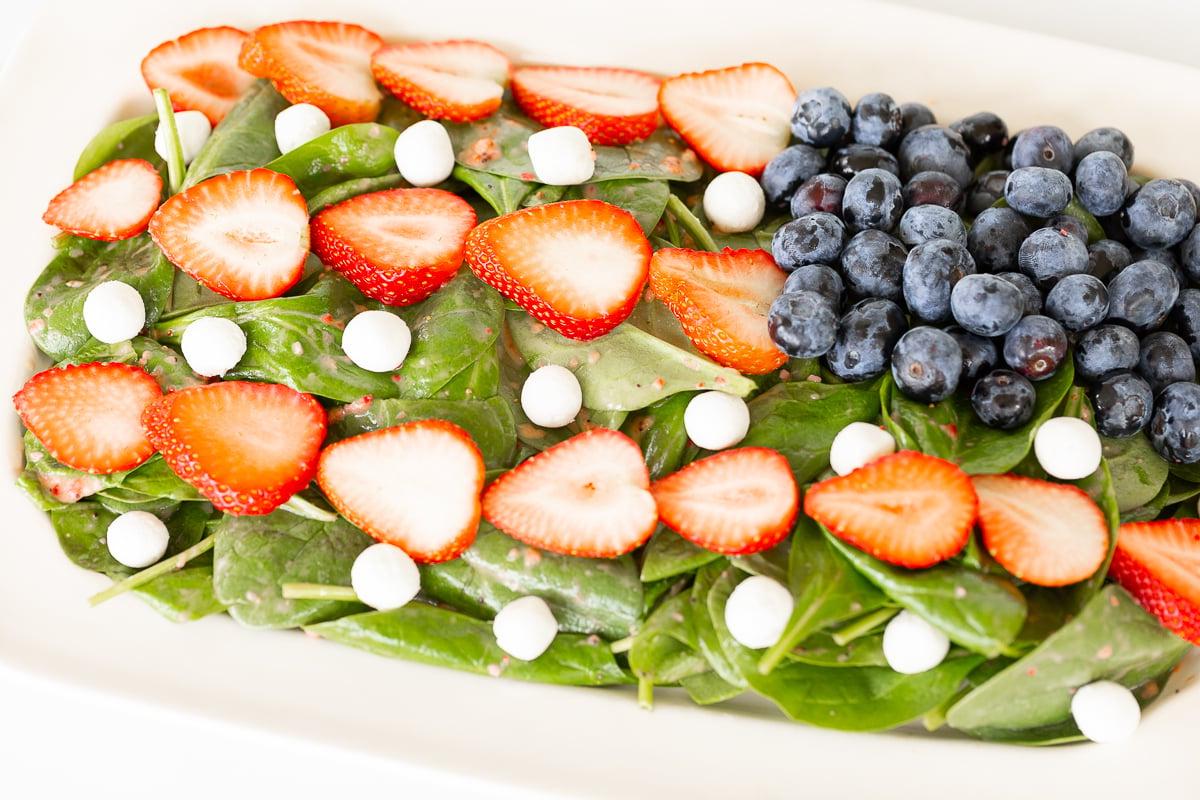 Grillbeilagen 5 Ein frischer Spinatsalat, belegt mit geschnittenen Erdbeeren, ganzen Heidelbeeren und weißen Käsekugeln, serviert in einer roten Schale.