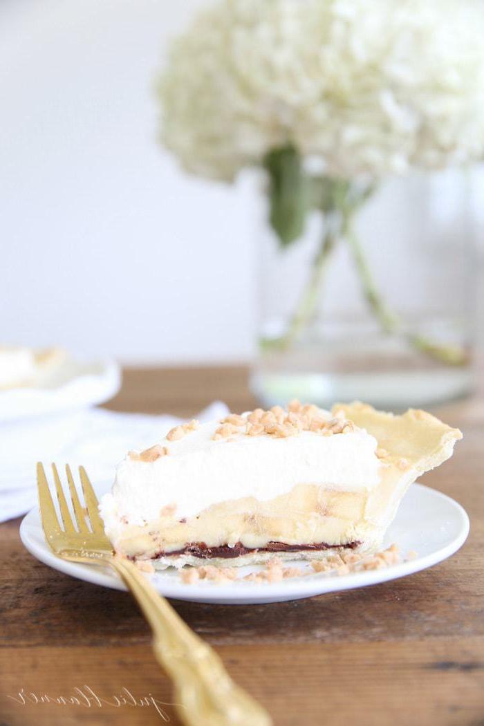 Bananenrezepte 6 Ein Stück einfacher Bananencremetorte auf einem weißen Teller mit goldener Gabel und weißen Blumen in einer Glasvase im Hintergrund.
