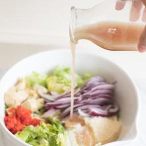 Rotweinessig-Vinaigrette 6 Eine Hand gießt Rotweinvinaigrette aus einer Glasflasche in eine große weiße Schüssel mit gehacktem Gemüse, darunter Salat, rote Zwiebeln und rote Paprika.