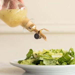 Weißwein-Vinaigrette 6 Eine Person gießt Weißweinessig-Vinaigrette aus einer Glasflasche mit Messingausgießer auf einen Teller mit frischem grünem Salat.