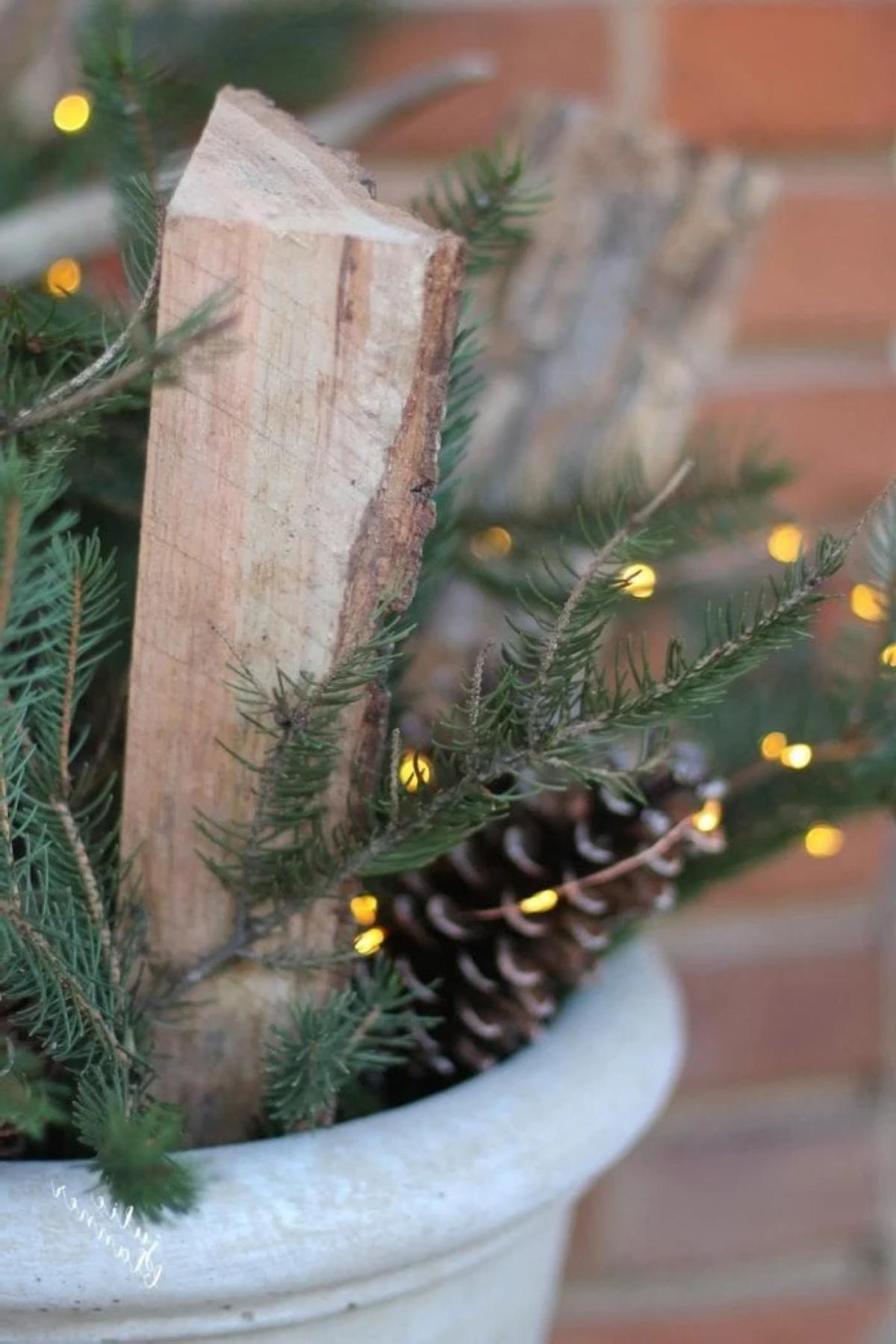 Ein Winterkübel für die Veranda mit geschnittenem Brennholz, immergrünen Pflanzen, Kiefernzapfen und Lichterketten.