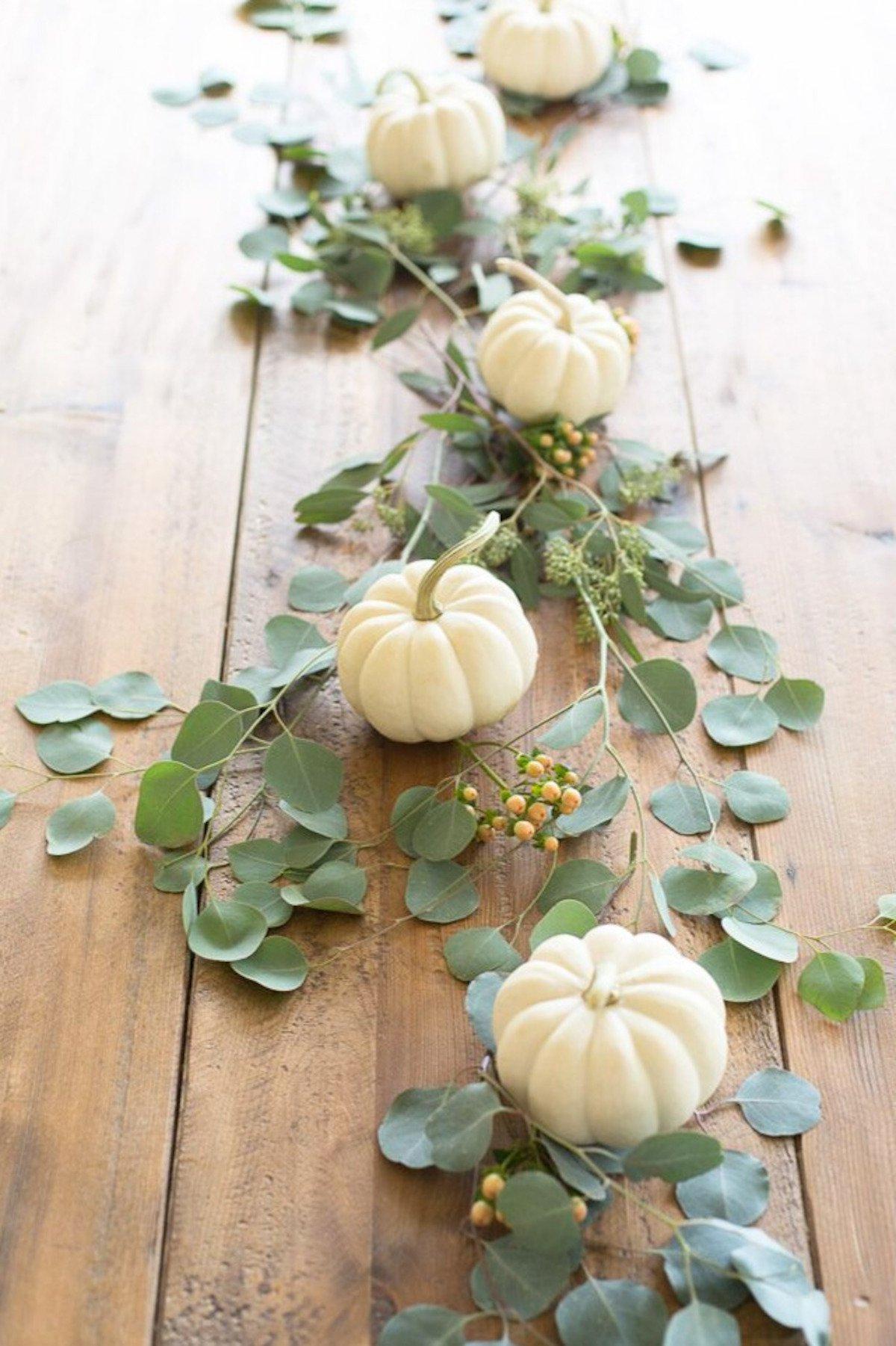 Ein herbstliches Mittelstück aus kleinen weißen Kürbissen und Eukalyptus in der Mitte eines Holztisches.