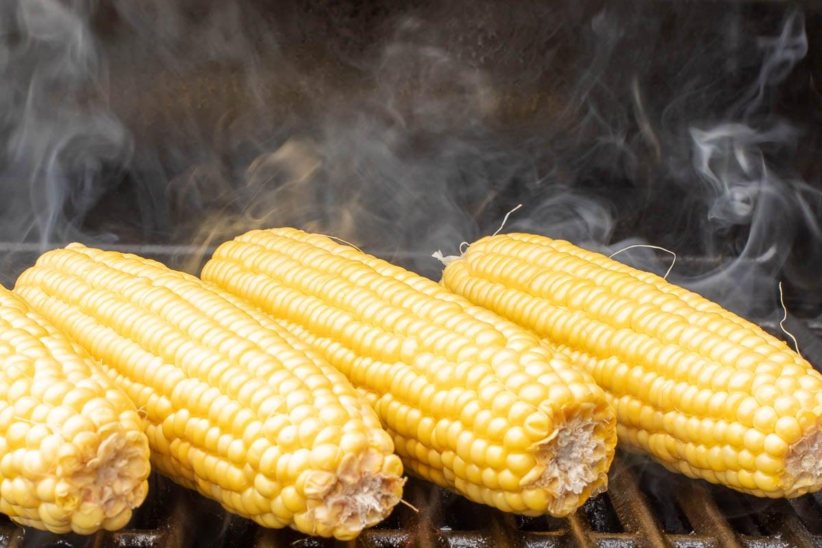 Gegrillter Maiskolben 7 Gegrillter Süßmaiskolben, der auf einem Grillrost gegrillt wird, mit Dampf, der von der heißen Oberfläche aufsteigt.