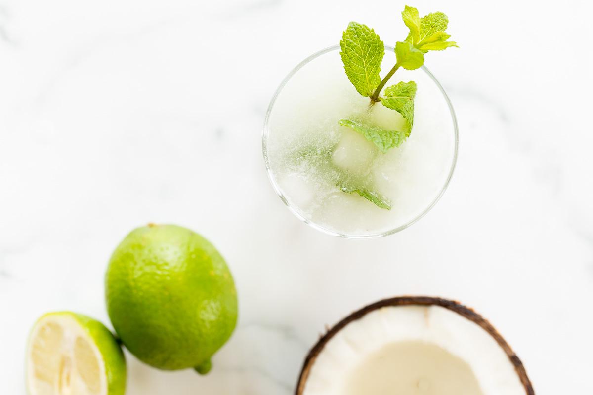 Kokosnuss-Mojito 8 Ein Kokos-Mojito, garniert mit Minze, auf einer Marmorfläche. Limetten und eine offene Kokosnuss daneben.